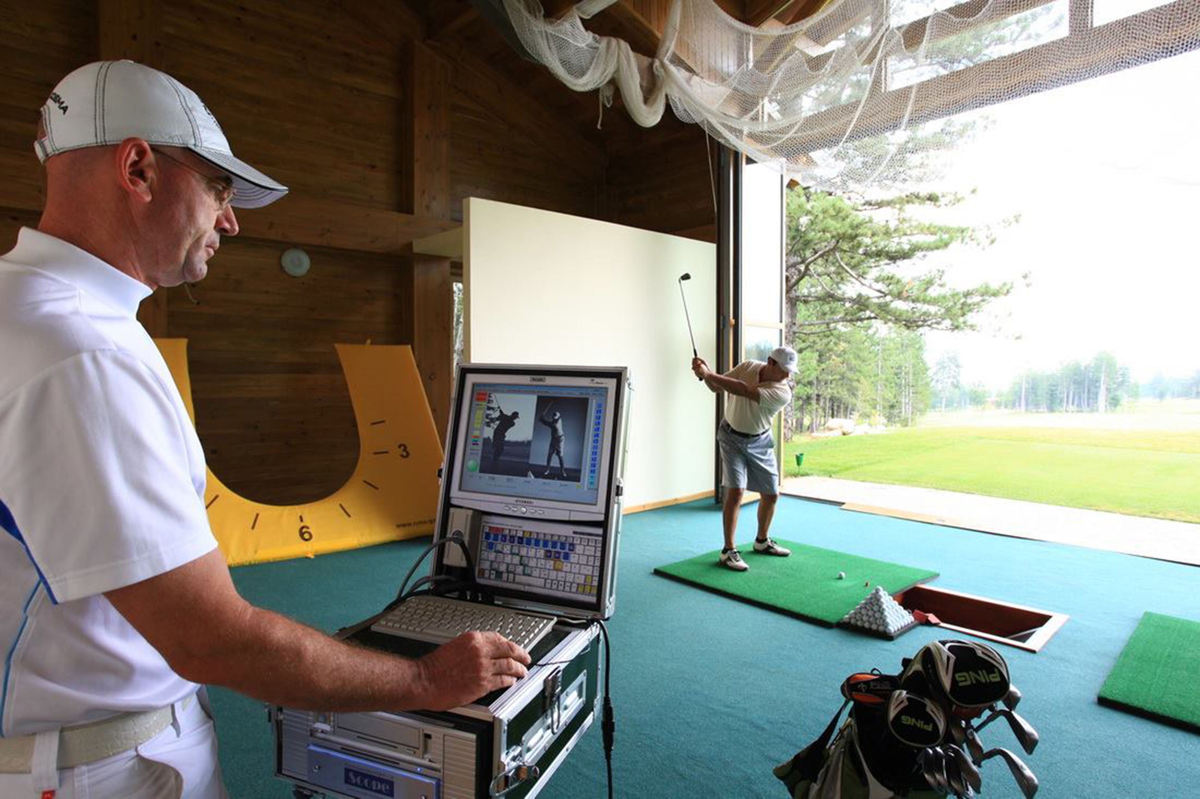 Un hombre usando una computadora portátil en una habitación con una pantalla grande mostrando un balanceo de golf, un bolso de golf y un tapete de golf.