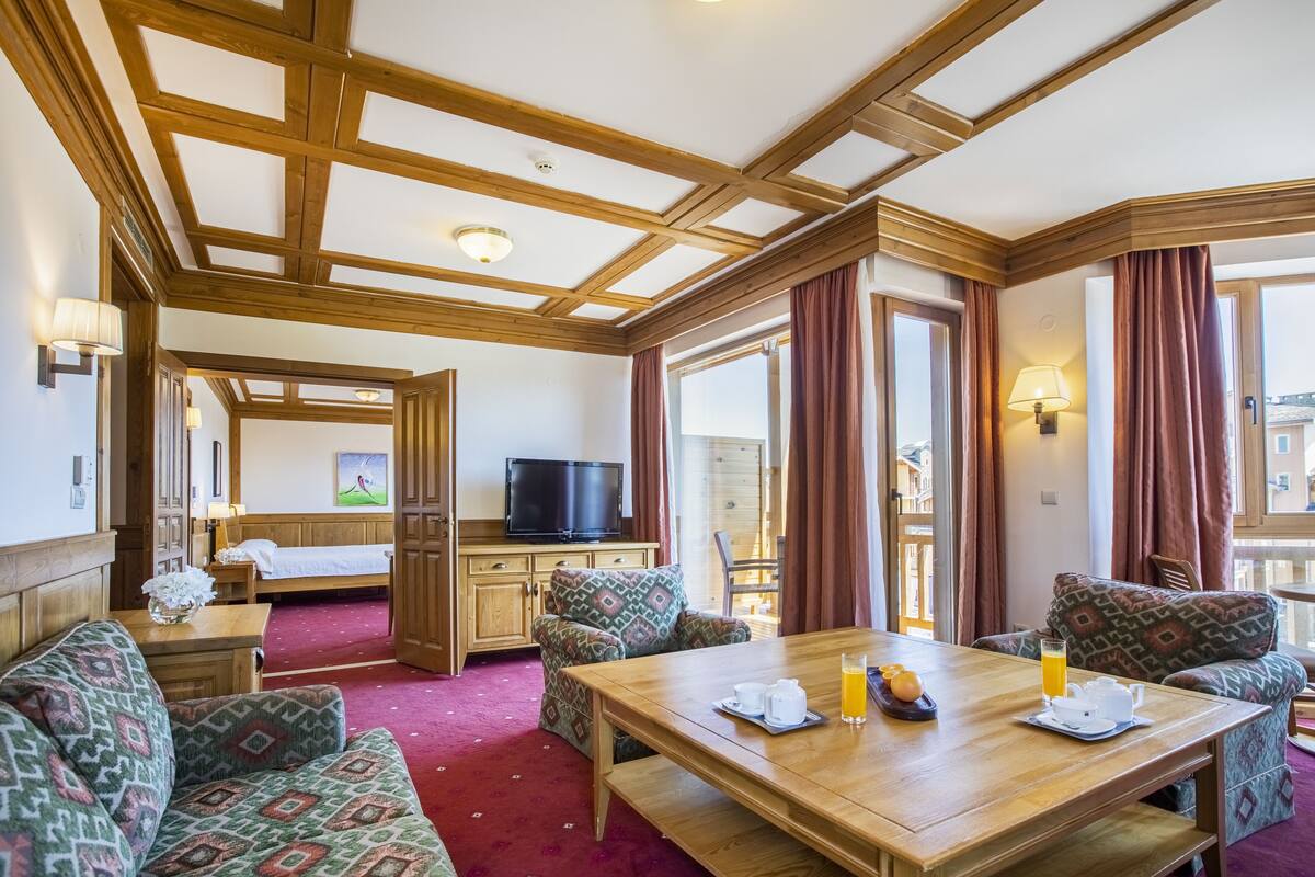 A hotel room with a wooden ceiling, a TV, a bed, a dining table with breakfast items, and large windows with curtains.