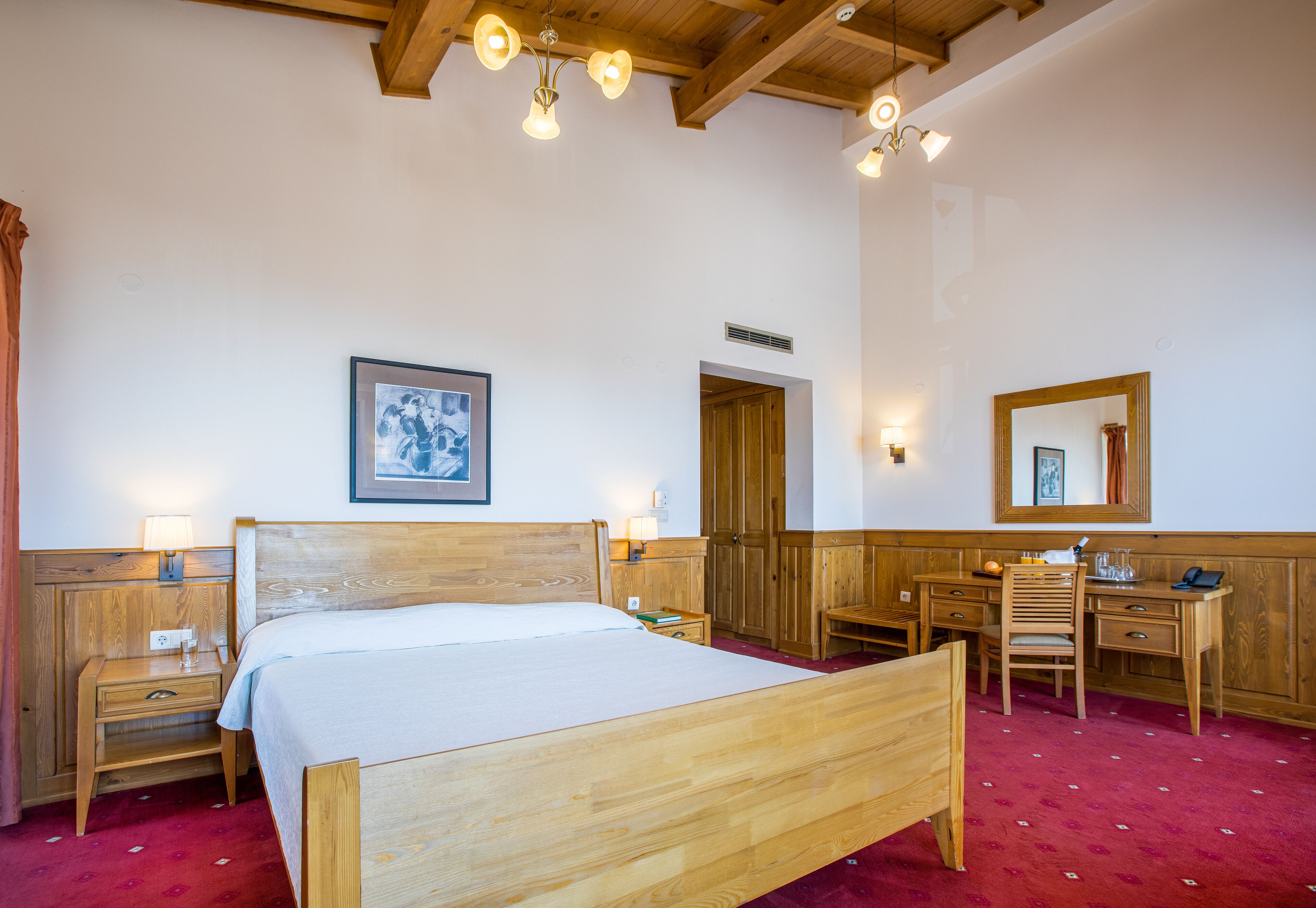 Una cama de madera con colchón blanco, una mesita de noche de madera con una lámpara, un escritorio de madera con una silla, un espejo y un cuadro en la pared.