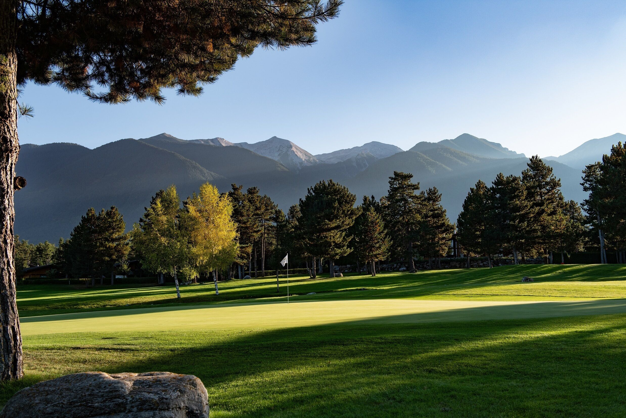 A golf course with a flag, trees, and mountains in the background.