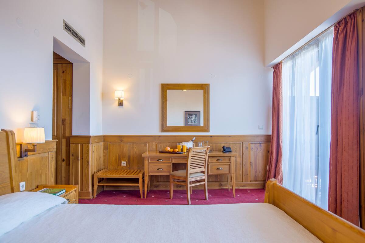 A hotel room with a bed, wooden desk, chair, and a large window with curtains.