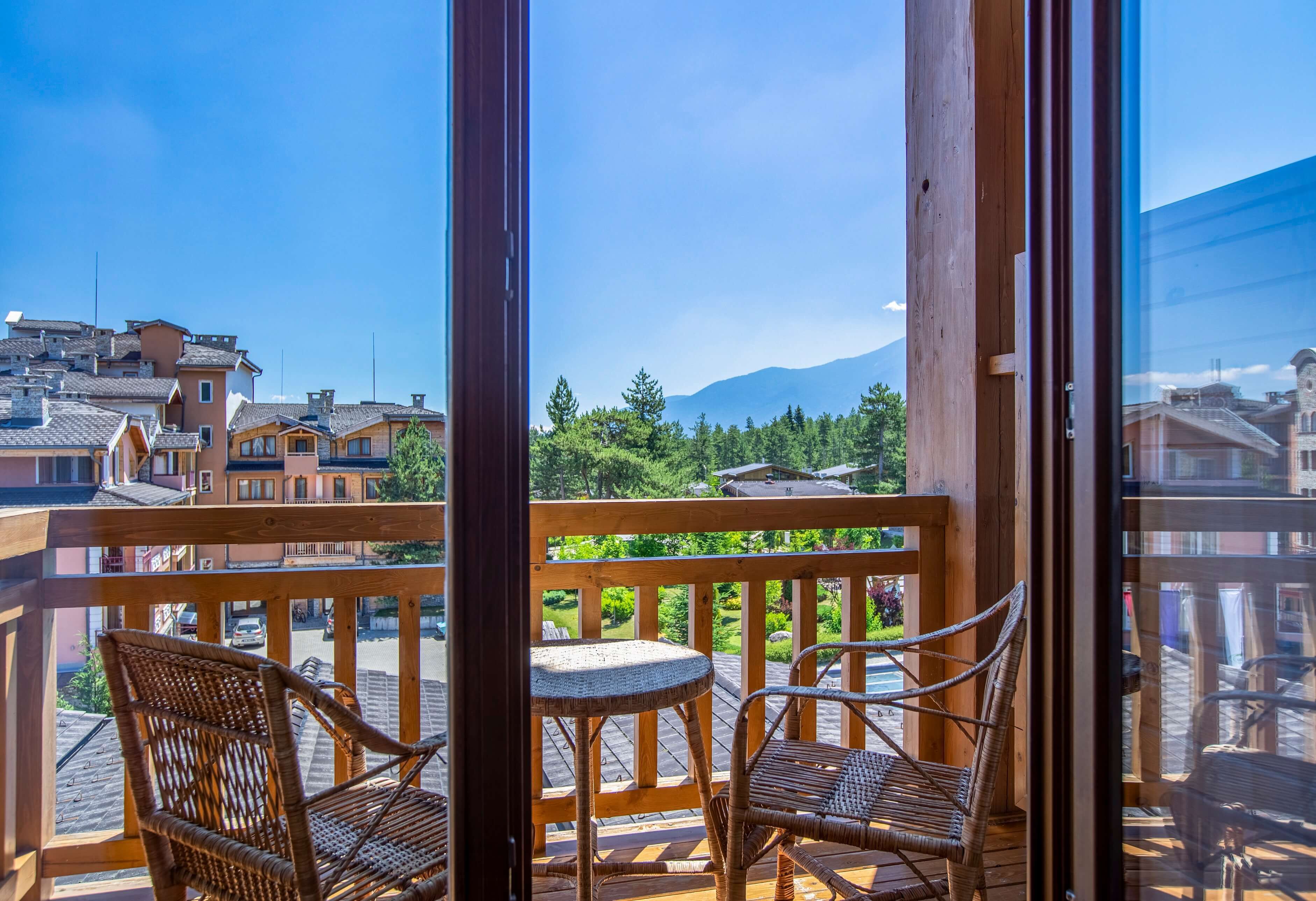 A balcony with wicker chairs and a small table, overlooking a residential area with houses and a mountain in the background.