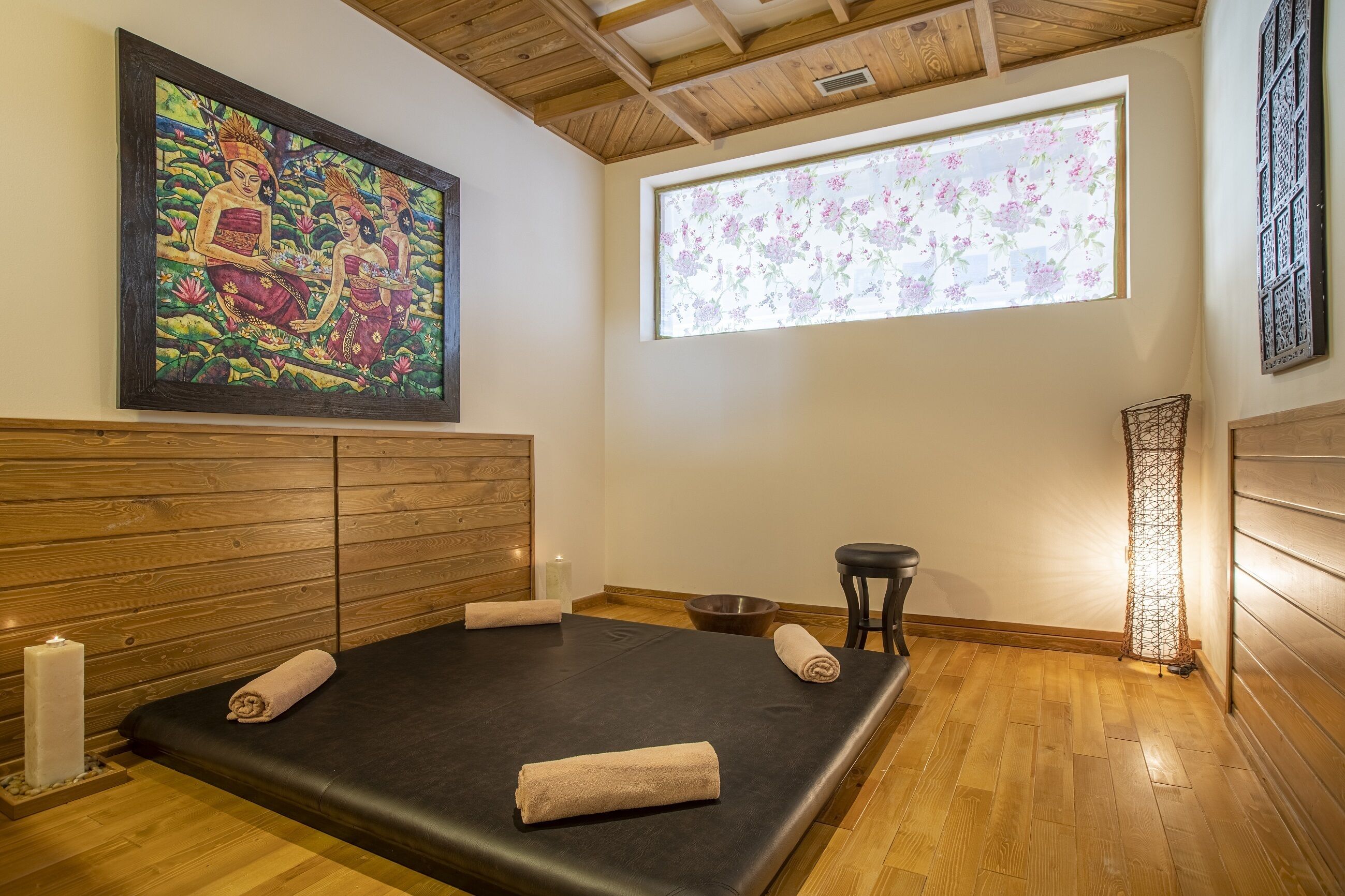 A room with a wooden ceiling, a large painting, a window with floral blinds, a massage table with towels, a candle, and a small stool.