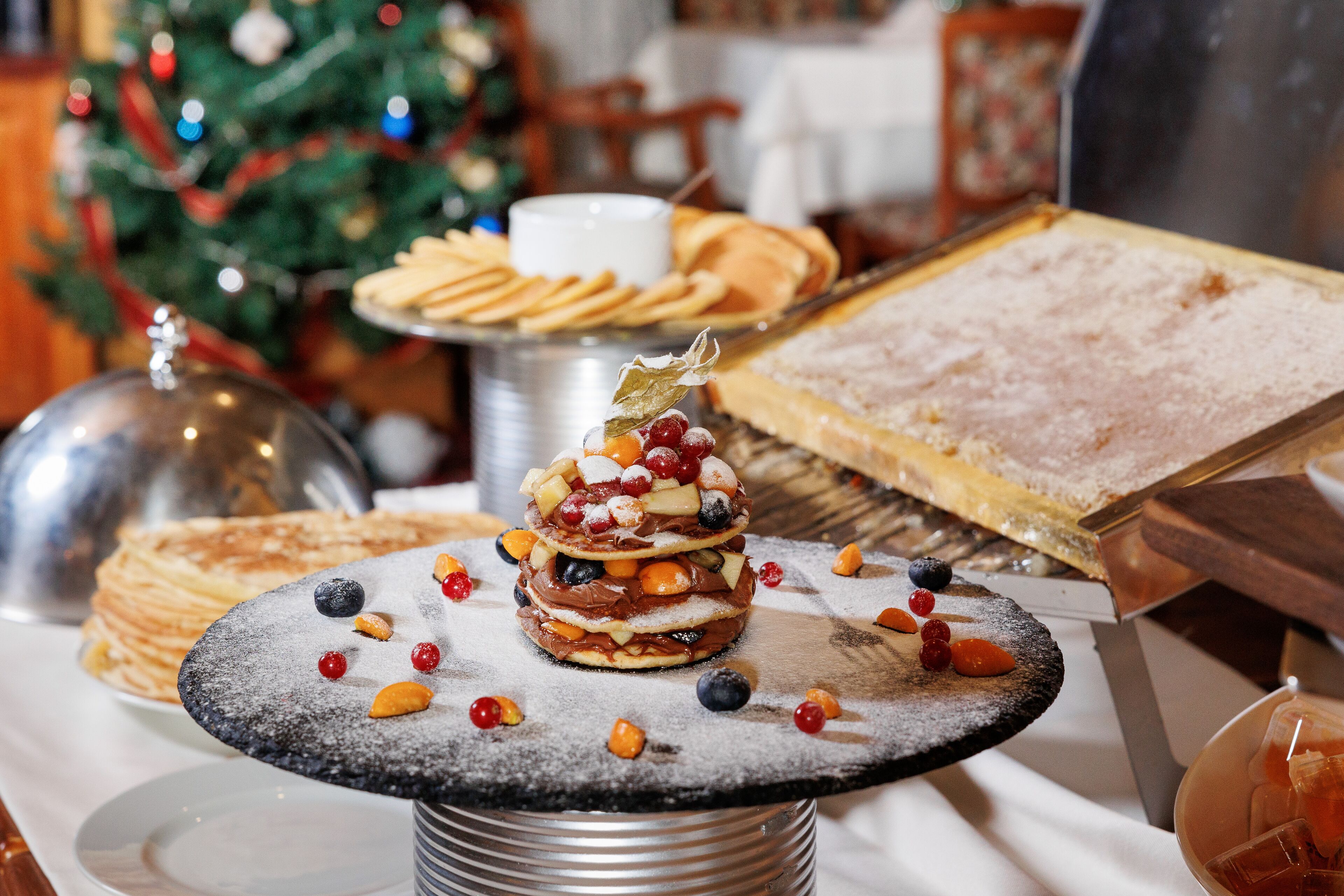Un bufé con diversos postres y pasteles, incluyendo un pastel en capas con frutas y nueces, y un pastel de chocolate con azúcar glas.