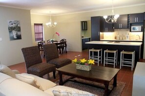 A living room with a sofa, two armchairs, a coffee table with flowers, a dining area with a table and chairs, and a kitchen with dark cabinets and a white countertop.
