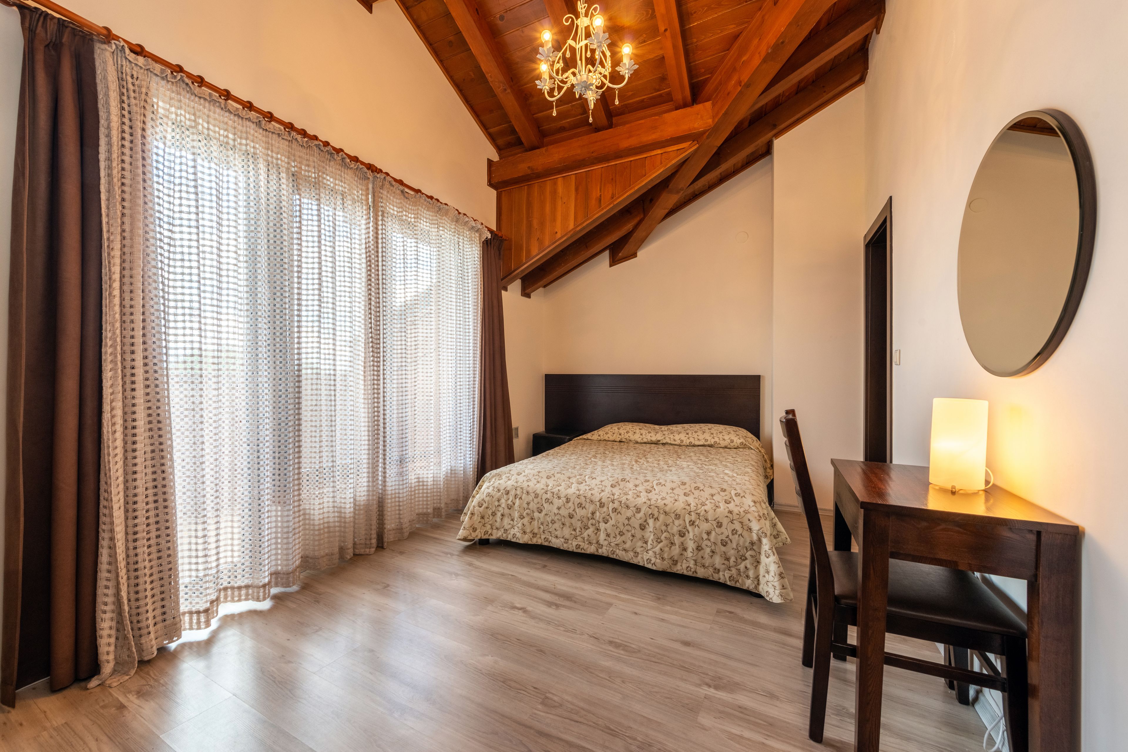 Een slaapkamer met een houten plafond, een bed met een bloemenprint deken, een bureau met een lamp en een grote spiegel.
