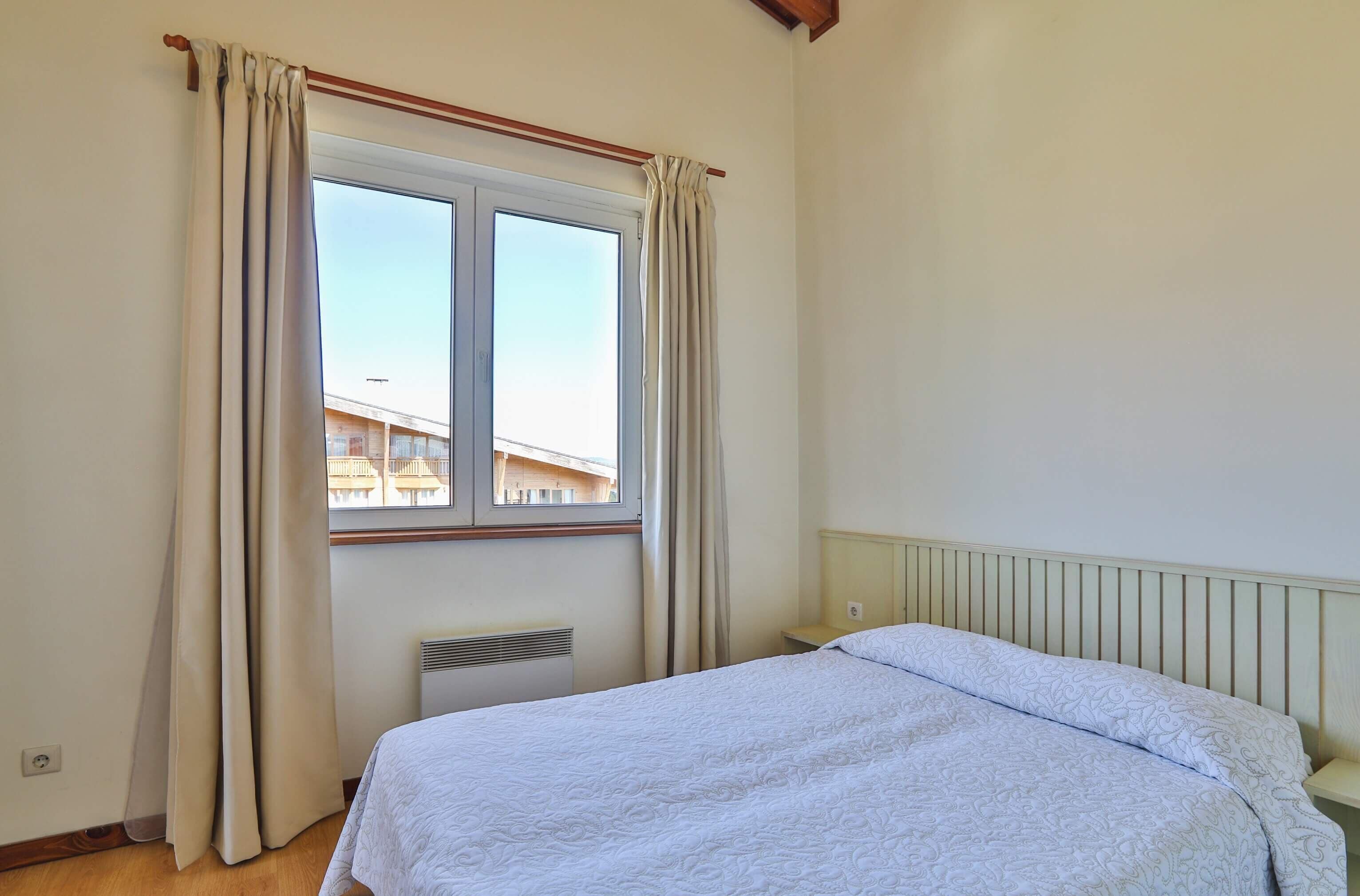 A bedroom with a bed, a window with curtains, a radiator, and a view of buildings outside.
