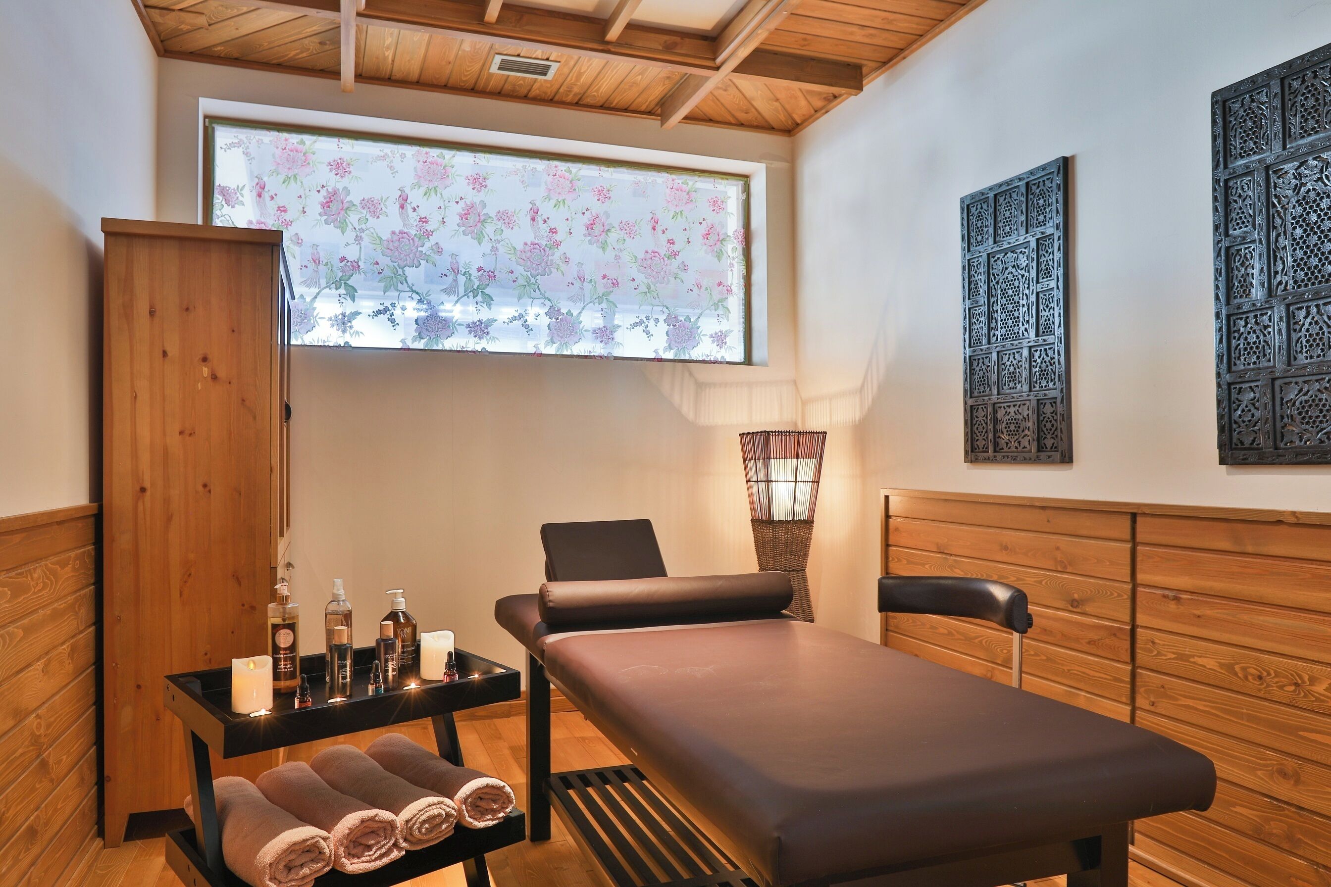 Un cuarto de spa con camilla de masaje, paneles de madera, una ventana con persianas florales y una lámpara fijada a la pared.
