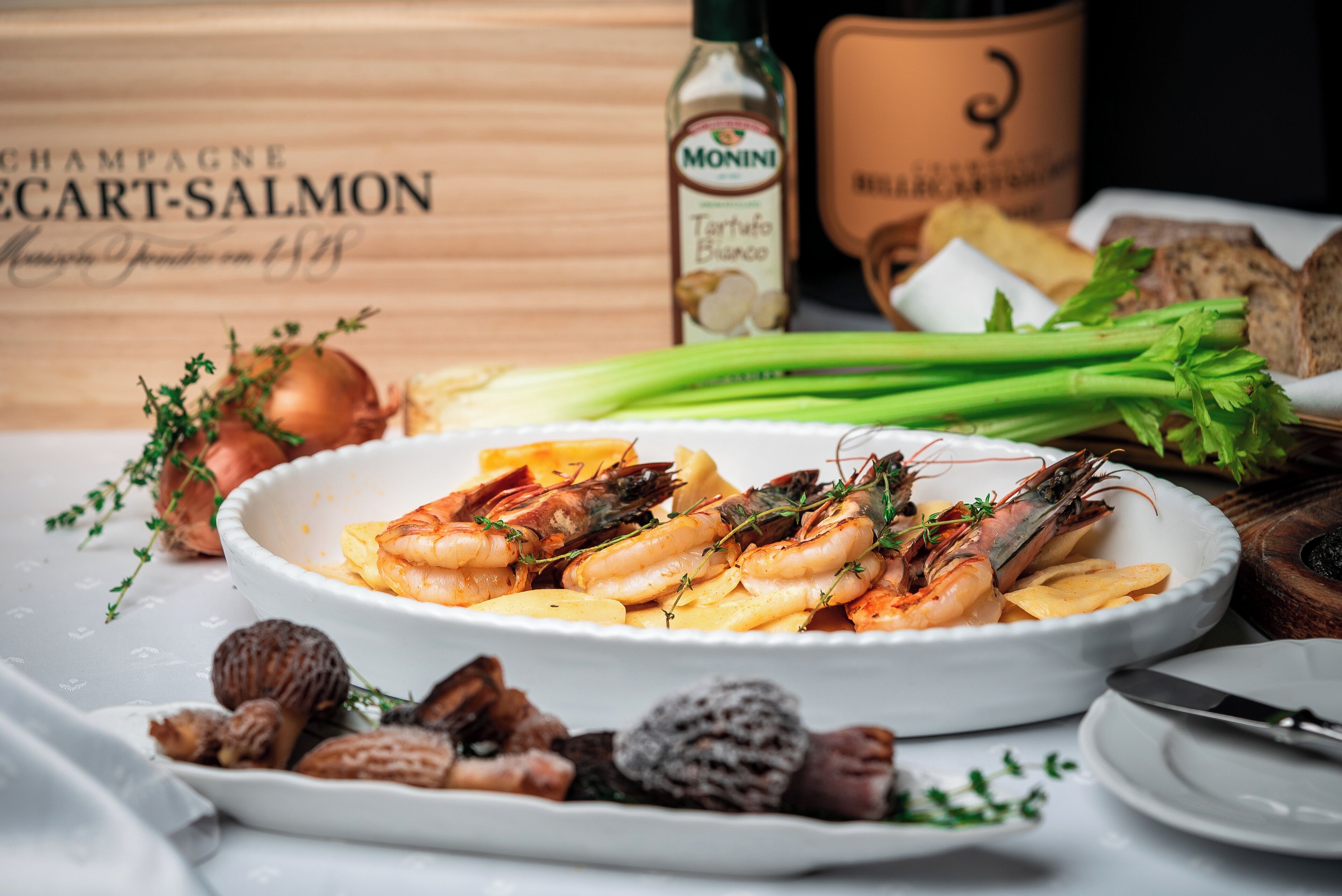 A plate of cooked shrimp with herbs, a bottle of Monini sauce, and some bread on a table.