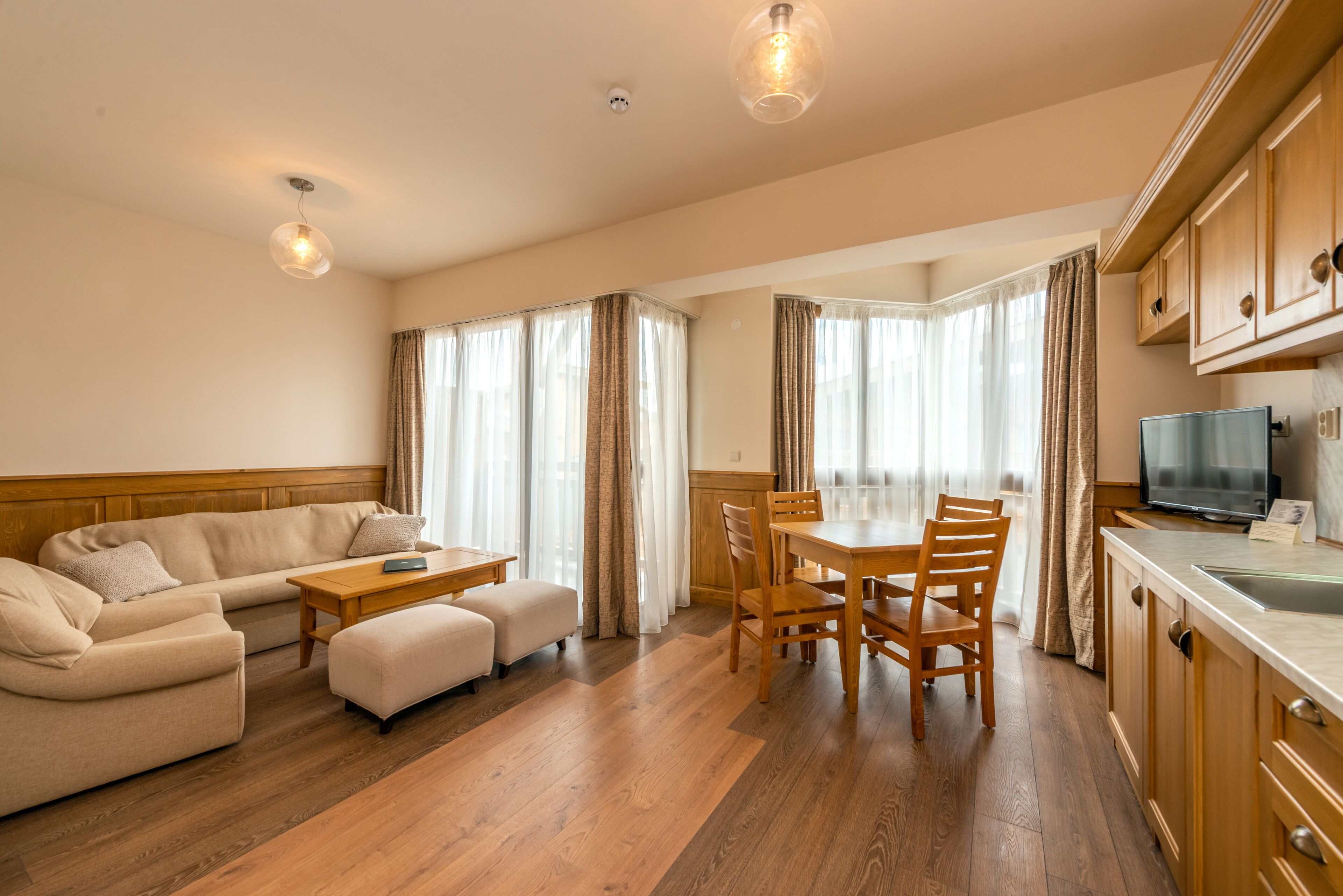 Un salon moderne avec un canapé, un pouf, une table à manger en bois et des chaises. On y trouve un téléviseur posé sur un meuble et une fenêtre avec des rideaux.