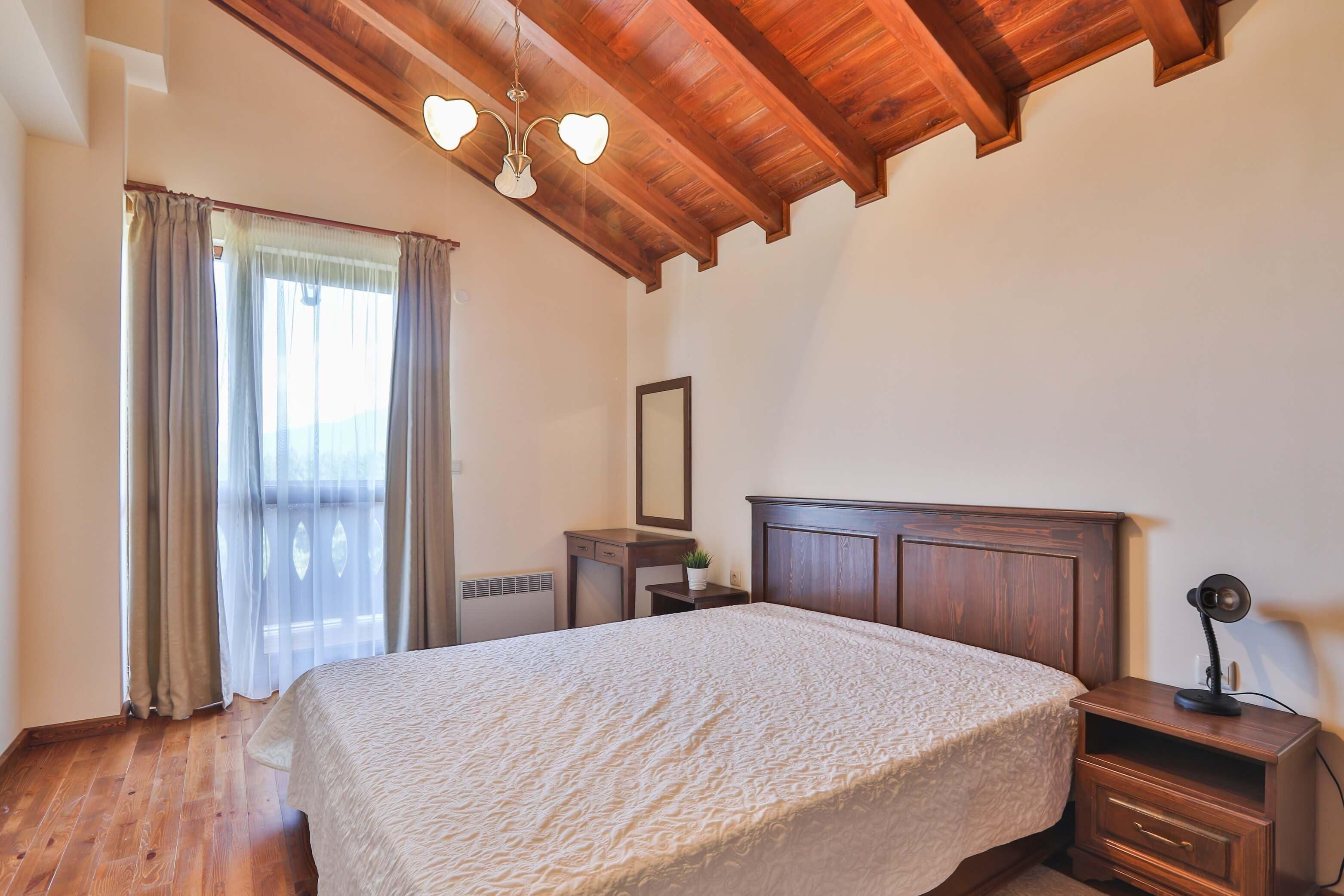 Un dormitorio con techo de madera, una cama con colcha blanca texturizada, una mesita de noche con lámpara y una ventana con cortinas.