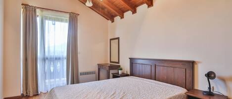 Un dormitorio con techo de madera, una cama con colcha blanca texturizada, una mesita de noche con lámpara y una ventana con cortinas.