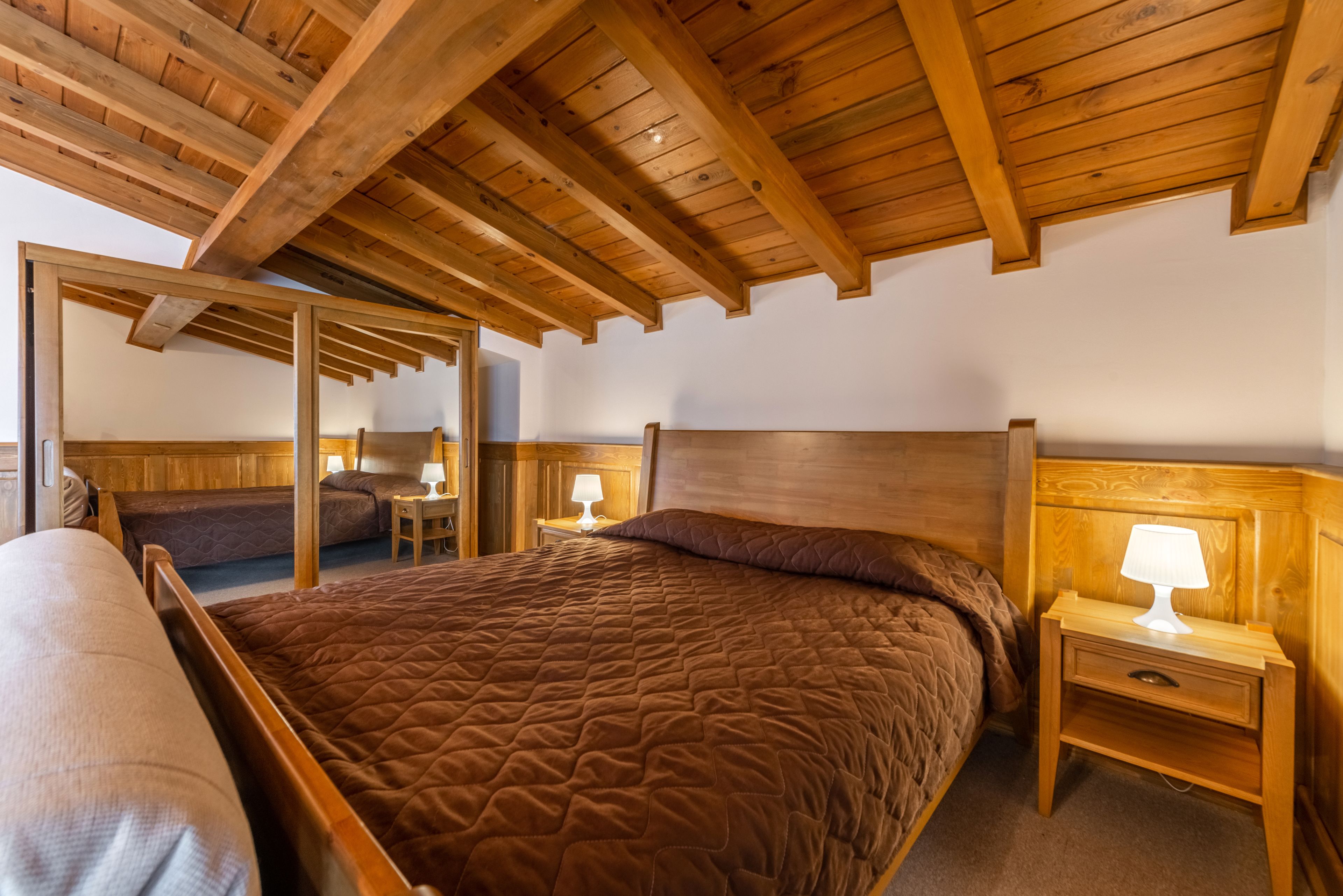 Une chambre à coucher avec un plafond en bois, un grand lit recouvert d’une courtepointe marron, deux tables de chevet avec des lampes et une tête de lit en bois.