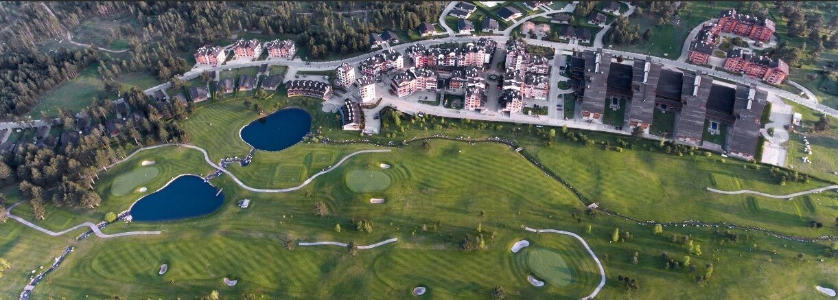 Vista aérea de un campo de golf con múltiples búnkeres, zonas de agua y una zona residencial con edificios y carreteras.