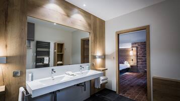 A modern bathroom with a wooden vanity, a large mirror, and a separate bedroom visible through an open door.