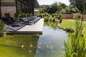 Ein Teich mit Koi-Karpfen, eine Terrassenfläche, umgeben von Grünpflanzen und einem Backsteinhaus.