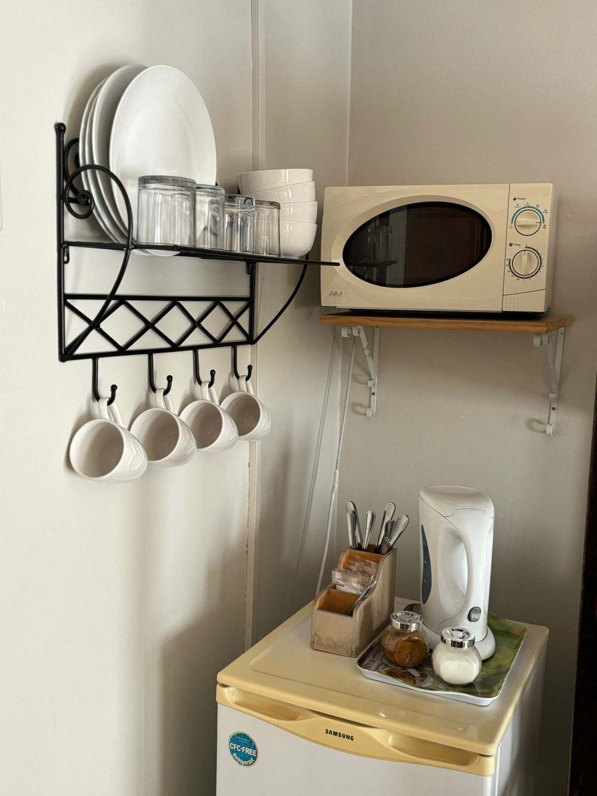 Una cocina con microondas, cafetera y estante con vajilla y tazas.