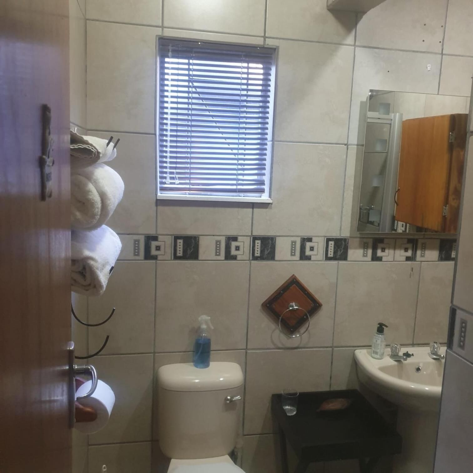 A bathroom with a toilet, sink, and window. There are towels hanging on the wall and a small table with a glass.