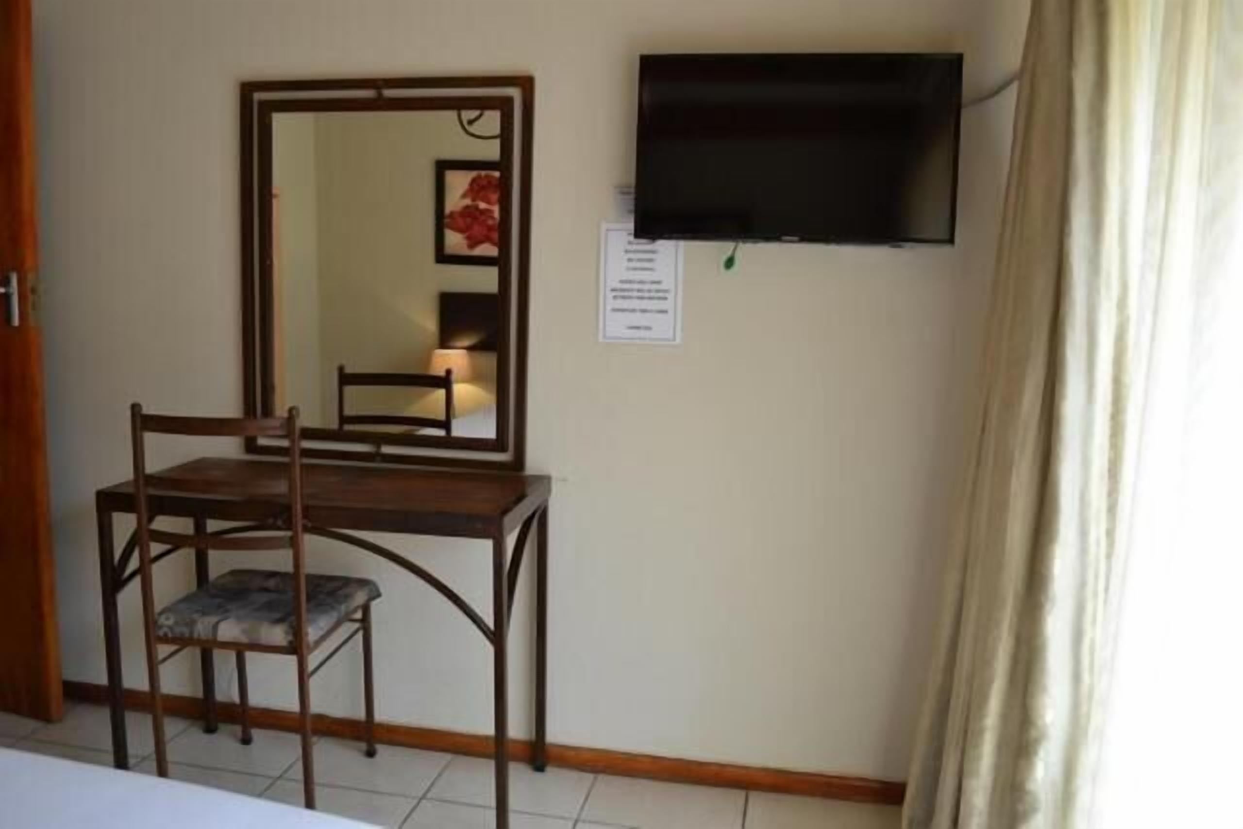 Una habitación con un escritorio de madera, una silla, un espejo, un televisor de pantalla plana y una cortina.