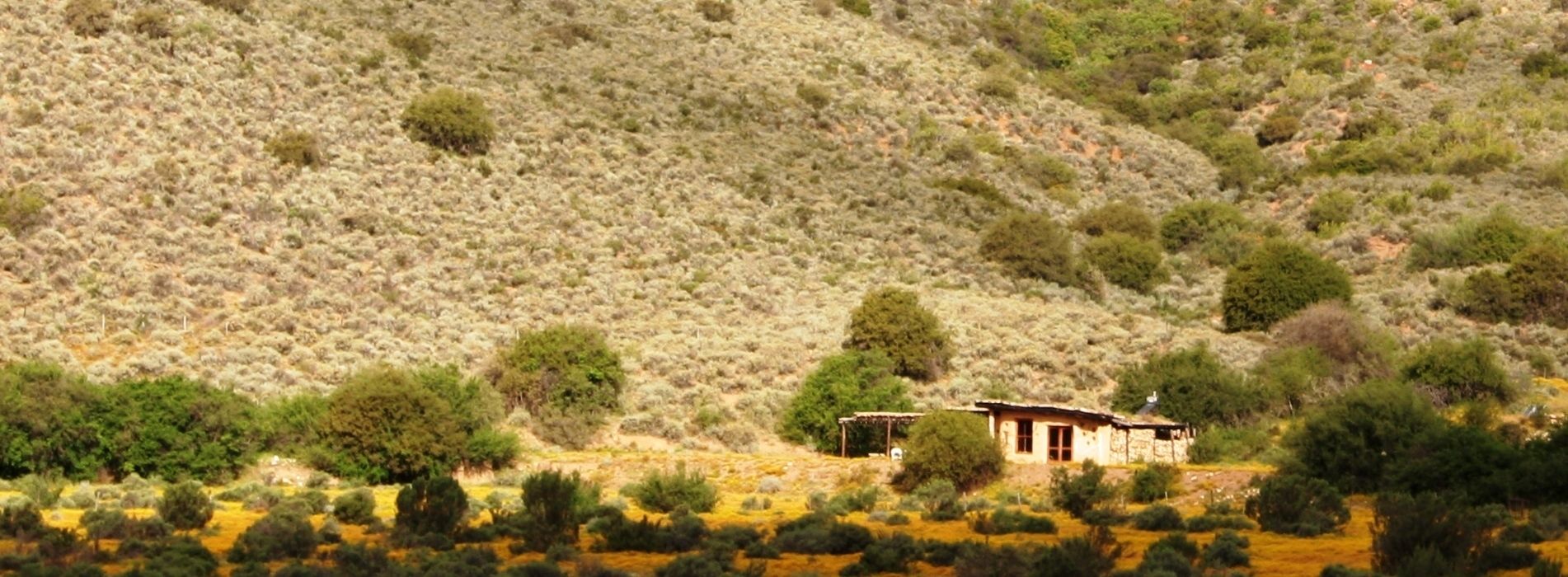 Una casa pequeña de construcción sencilla, rodeada de vegetación escasa y un paisaje montañoso.