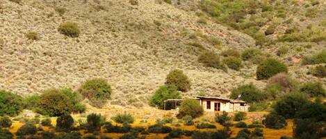 Una casa pequeña de construcción sencilla, rodeada de vegetación escasa y un paisaje montañoso.