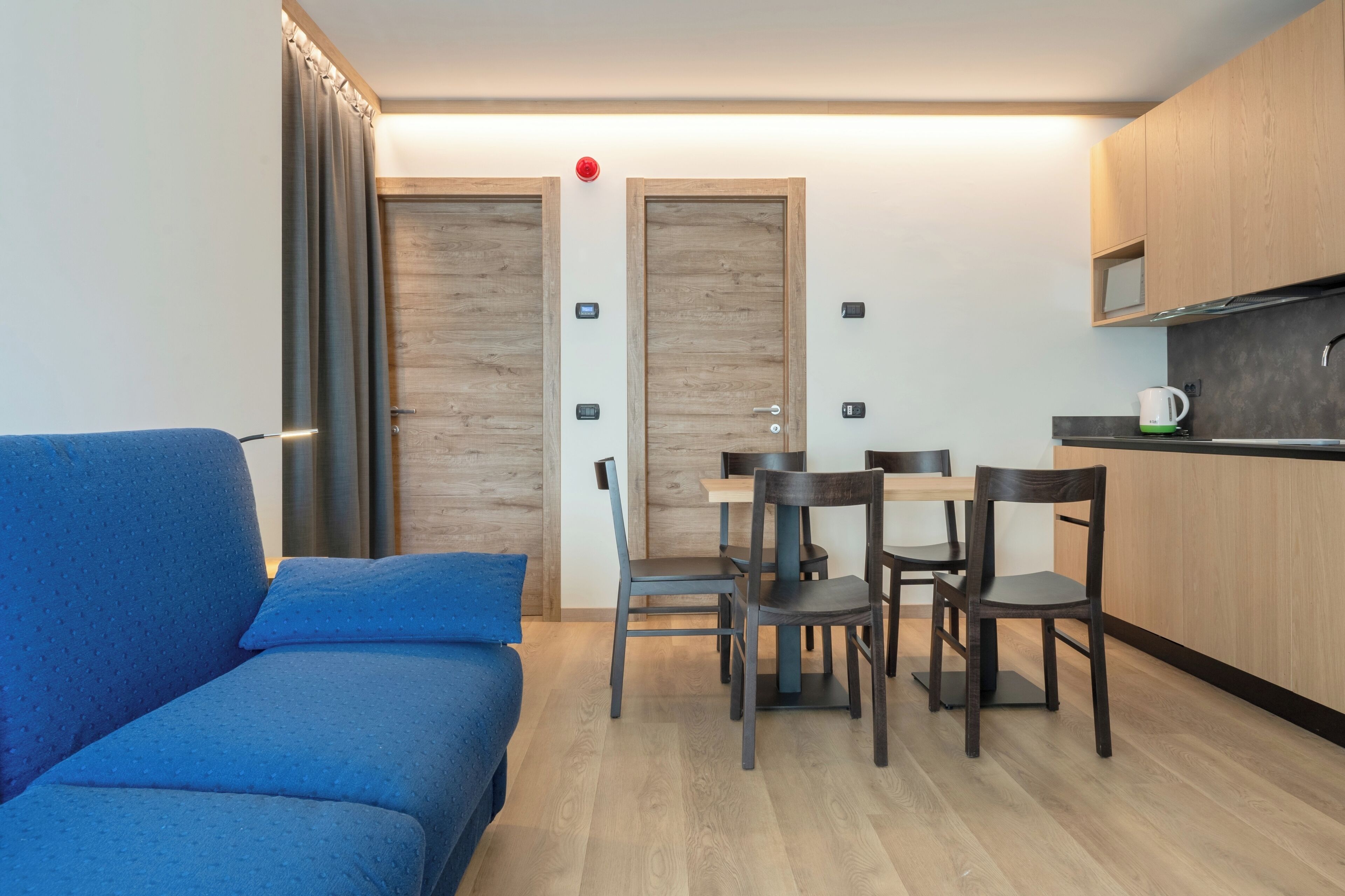A modern room with a blue sofa, a wooden dining table with four chairs, and a kitchenette with a microwave.