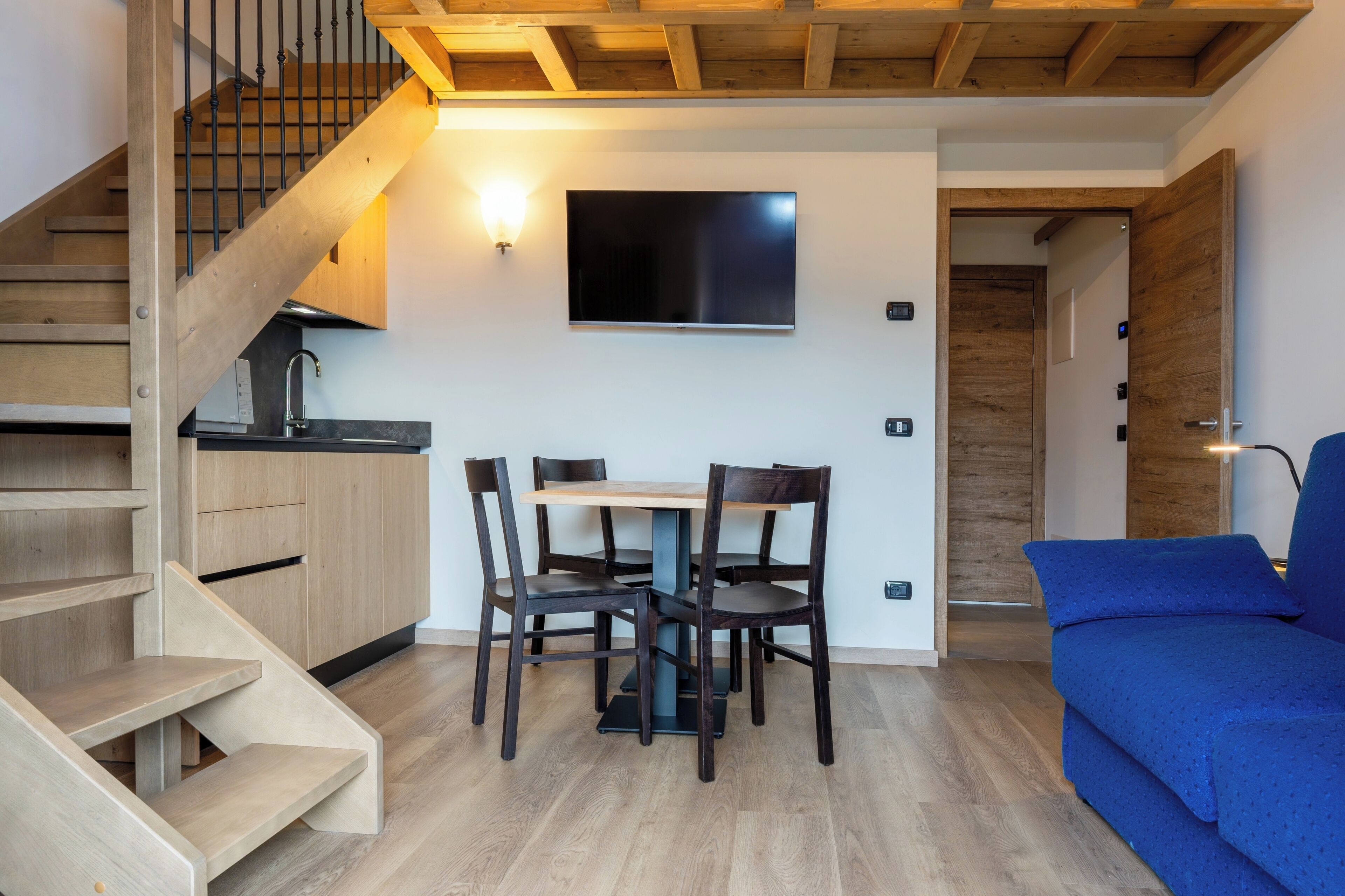 A modern living room with a wooden staircase, a dining area with a table and chairs, and a flat-screen TV mounted on the wall.