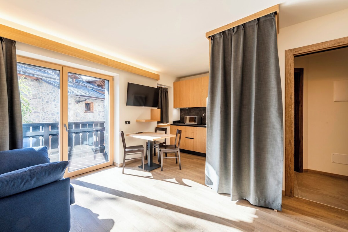 Una sala de estar moderna con un sofá, una mesa de comedor y sillas. Se observa una zona de cocina con armarios y un microondas. Una ventana grande con cortinas es visible, y una puerta da a otra habitación.