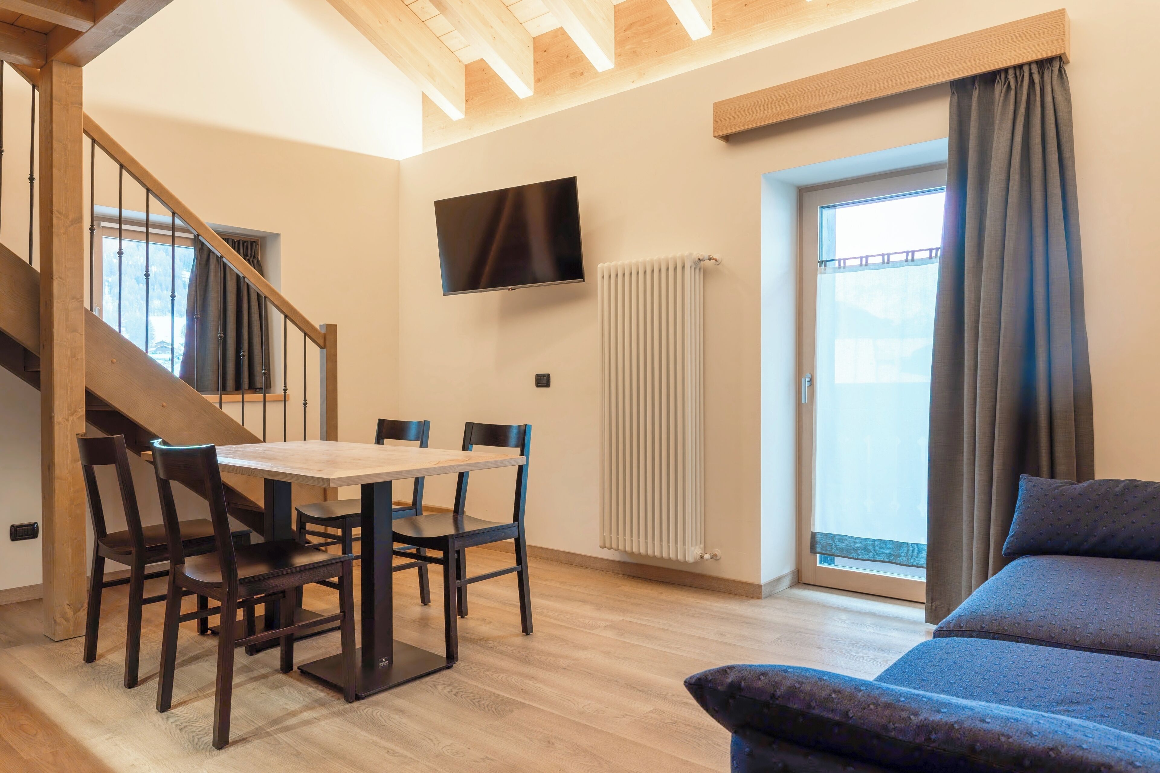 A modern living room with a wooden staircase, a dining area with a table and chairs, a flat-screen TV mounted on the wall, and a large window with curtains.