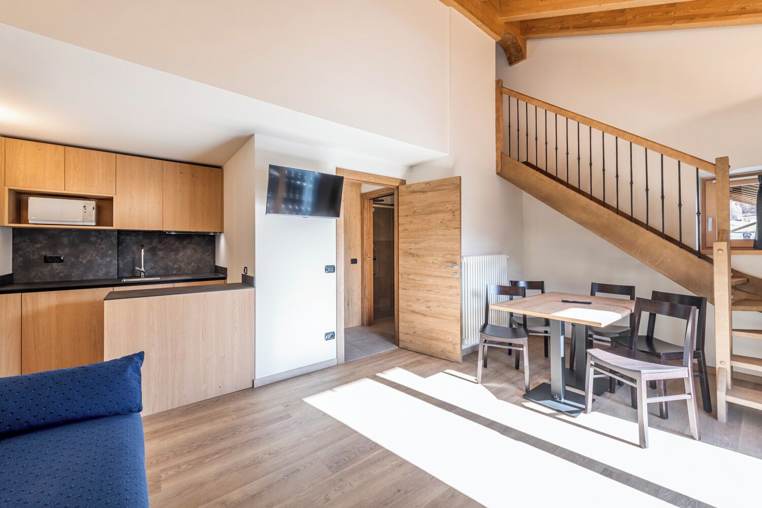 Una cocina moderna con armarios de madera y encimera negra, una zona de comedor con mesa y sillas, y una escalera de madera.