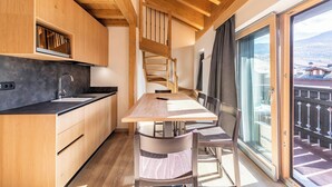 Una cocina moderna con armarios de madera, encimera negra y un comedor con mesa y sillas de madera. Una escalera de caracol conduce a un balcón con vistas a un paisaje montañoso.