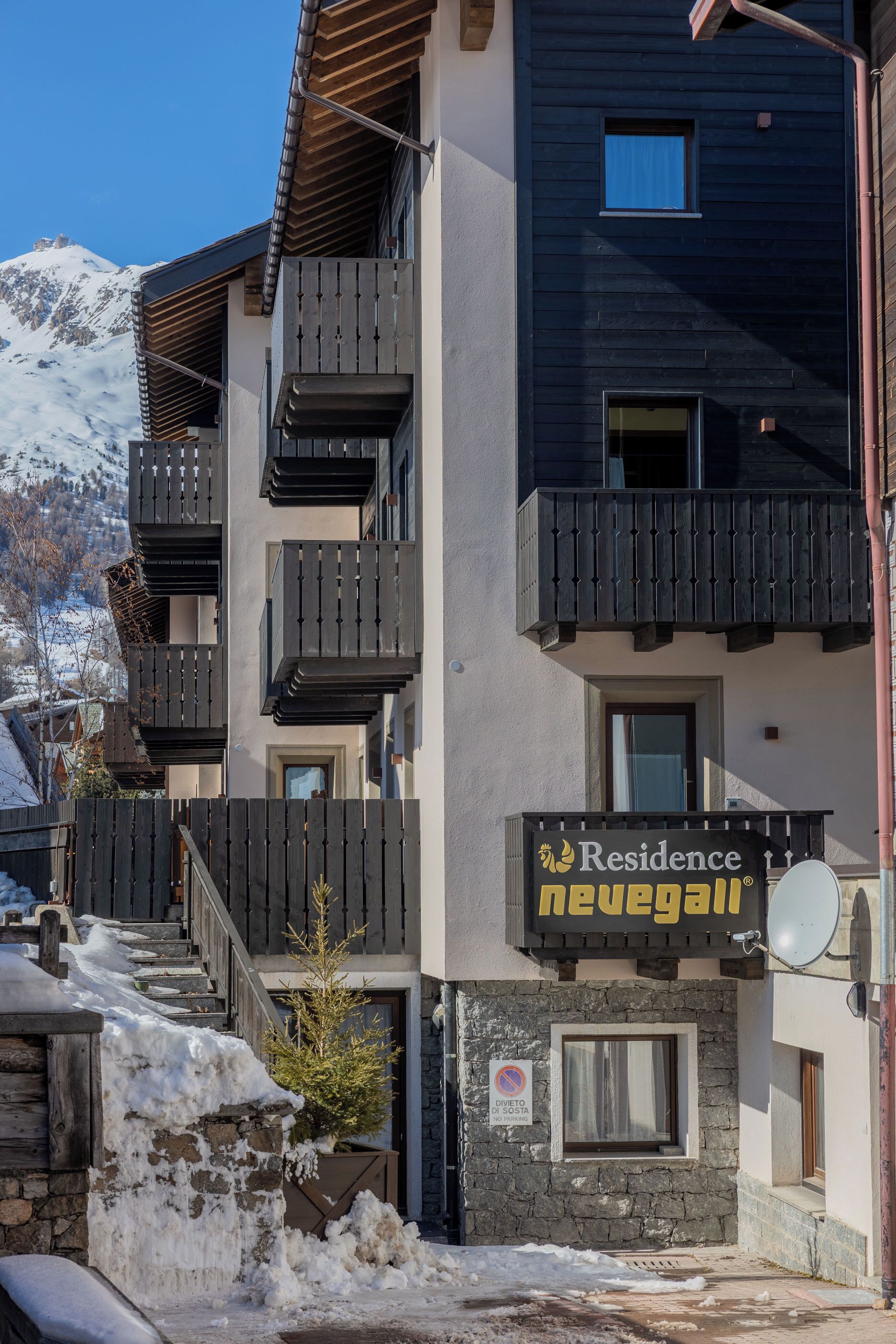 Un immeuble à plusieurs étages doté de balcons, une enseigne indiquant « Résidence Nevega », et une montagne enneigée en arrière-plan.