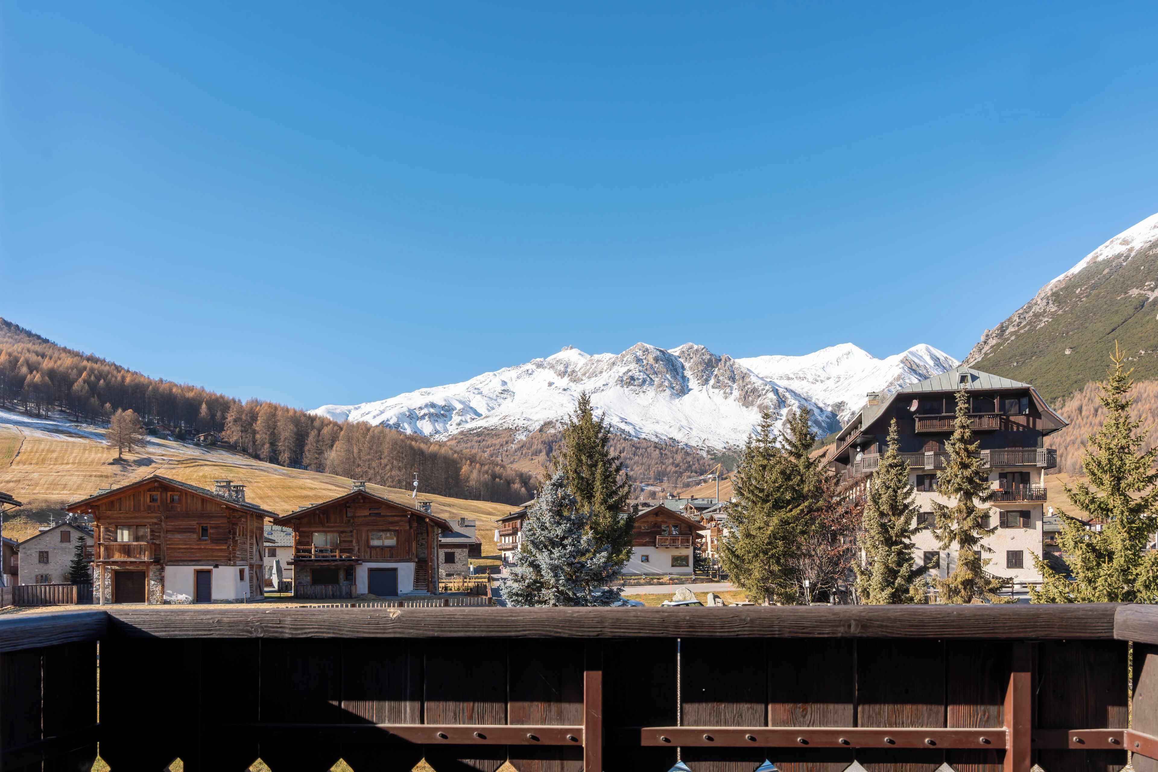 A wooden chalet with a mountain backdrop, featuring a wooden deck and a snow-covered mountain range.