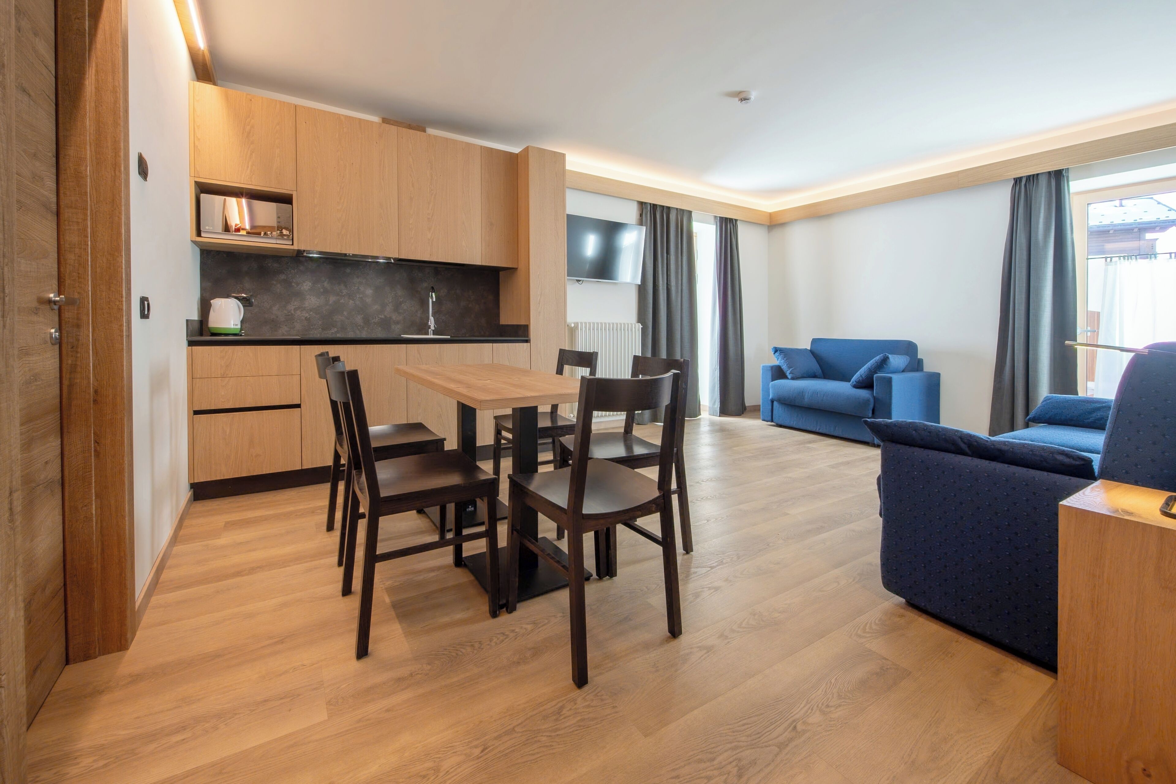 Un espacio de estar moderno con mesa de comedor de madera, sillas, un sofá azul y una zona de cocina con armarios de madera y encimera negra.
