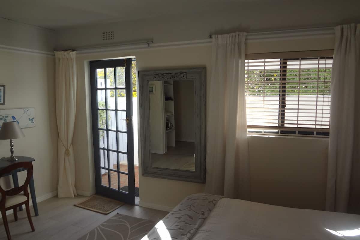 Um quarto com cama, uma cadeira, porta que dá para o exterior, janela com persianas e um espelho.