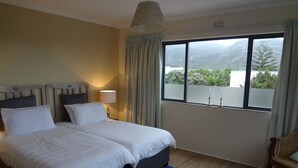 A hotel room with a bed, a nightstand, a lamp, a window with a view of mountains, and a chair. - Walker Bay Manor (Hermanus)