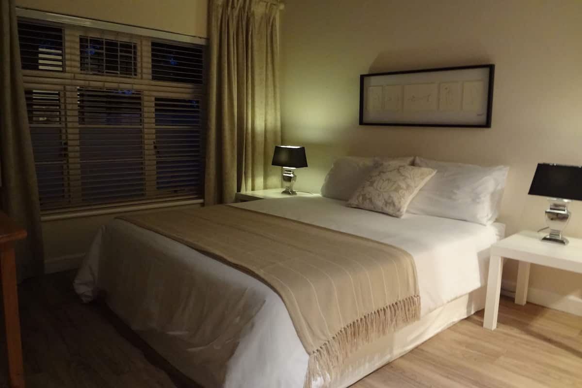 Uma cama bem arrumada com colcha bege, uma mesa de cabeceira com abajur e uma janela com persianas.