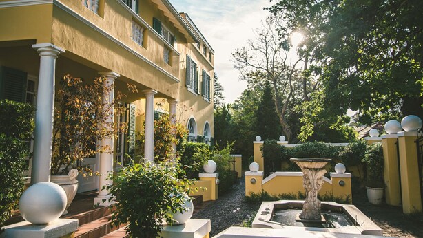Un cortile con una fontana, piante in vaso e un edificio giallo con colonne e persiane.