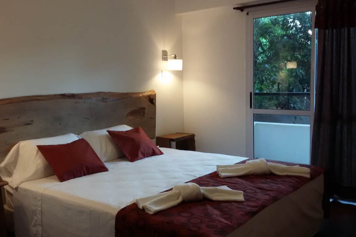 Une chambre d’hôtel avec un grand lit, deux tables de chevet, une tête de lit en bois et une fenêtre donnant sur un paysage verdoyant.