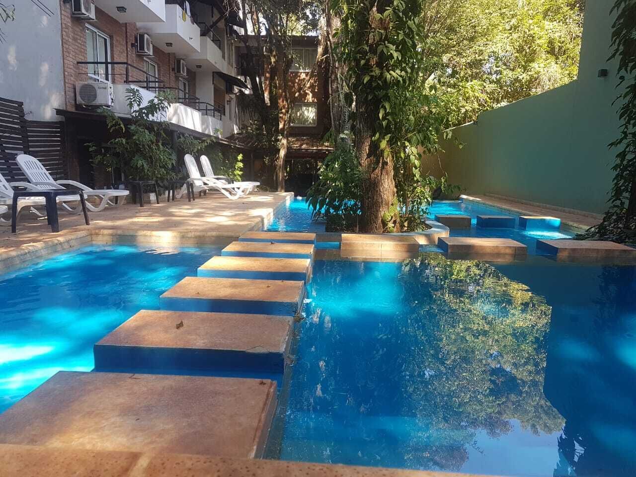 A swimming pool with steps leading into it, surrounded by lounge chairs and a building with multiple balconies.