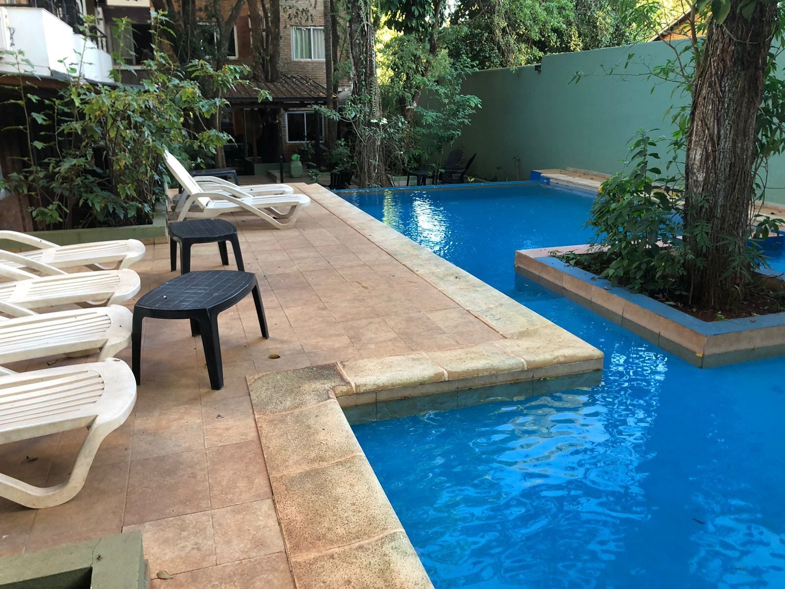A swimming pool with lounge chairs and a small table, surrounded by greenery and a building in the background.