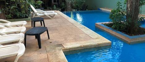 A swimming pool with lounge chairs and a small table, surrounded by greenery and a building in the background.