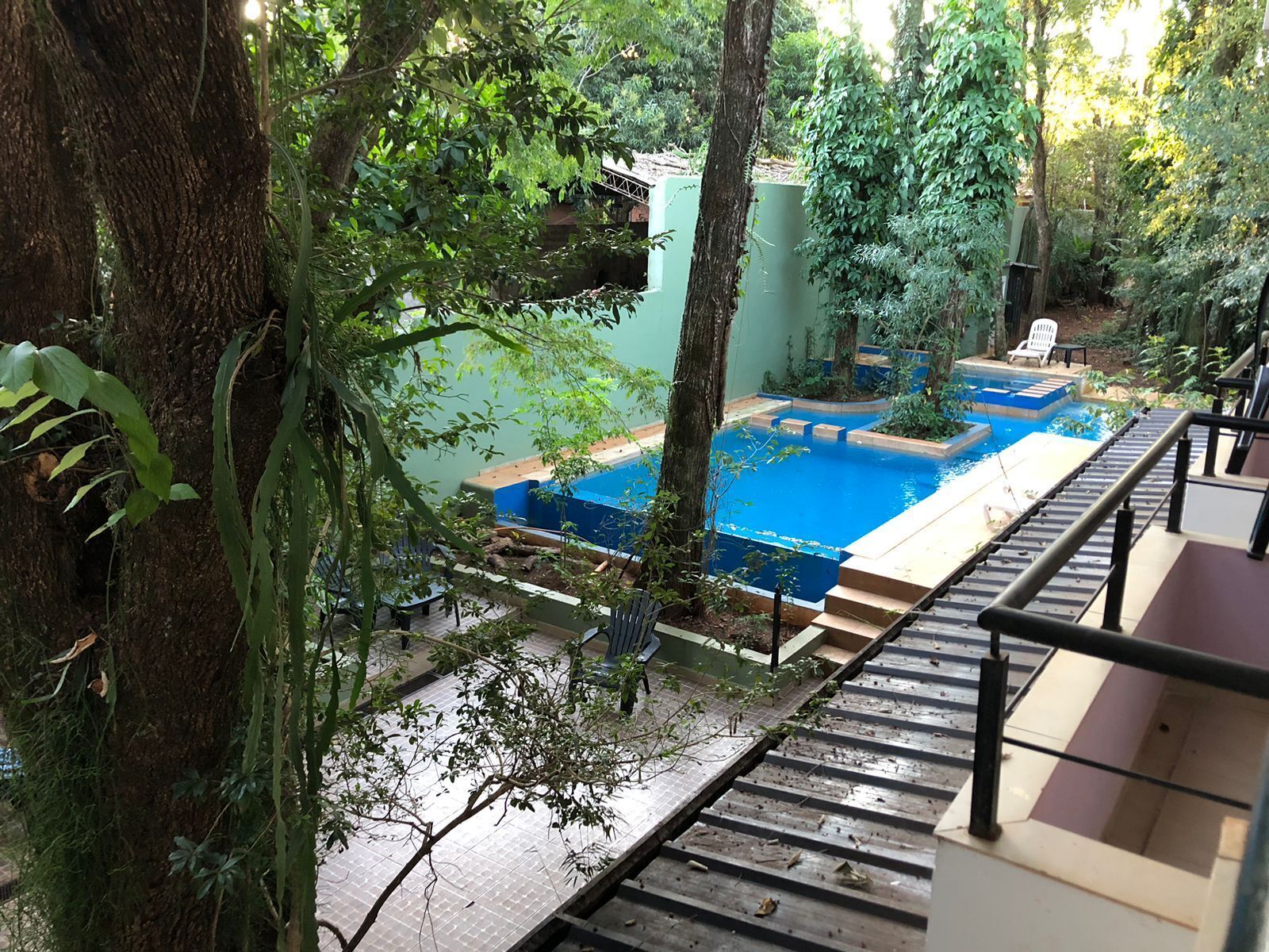 A pool area with a wooden deck and steps leading down to the water, surrounded by lush greenery and trees.