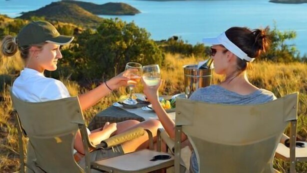 Two people sitting at an outdoor table with a view of the sea and hills in the background. - Otterskloof Private Game Reserve (Kopanong)