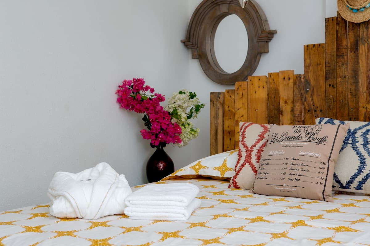 A neatly made bed with a patterned bedspread, a folded white towel, a vase with flowers, and a menu on the headboard.