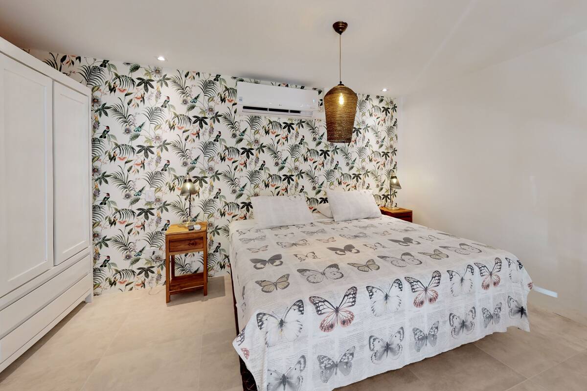 A bedroom with a floral wallpaper, a bed with a butterfly print bedspread, a wooden nightstand, a hanging lamp, and an air conditioning unit.