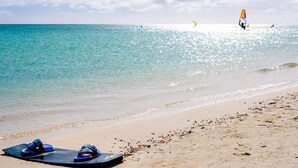 Una playa con agua turquesa cristalina, una orilla arenosa y una persona practicando windsurf a lo lejos.