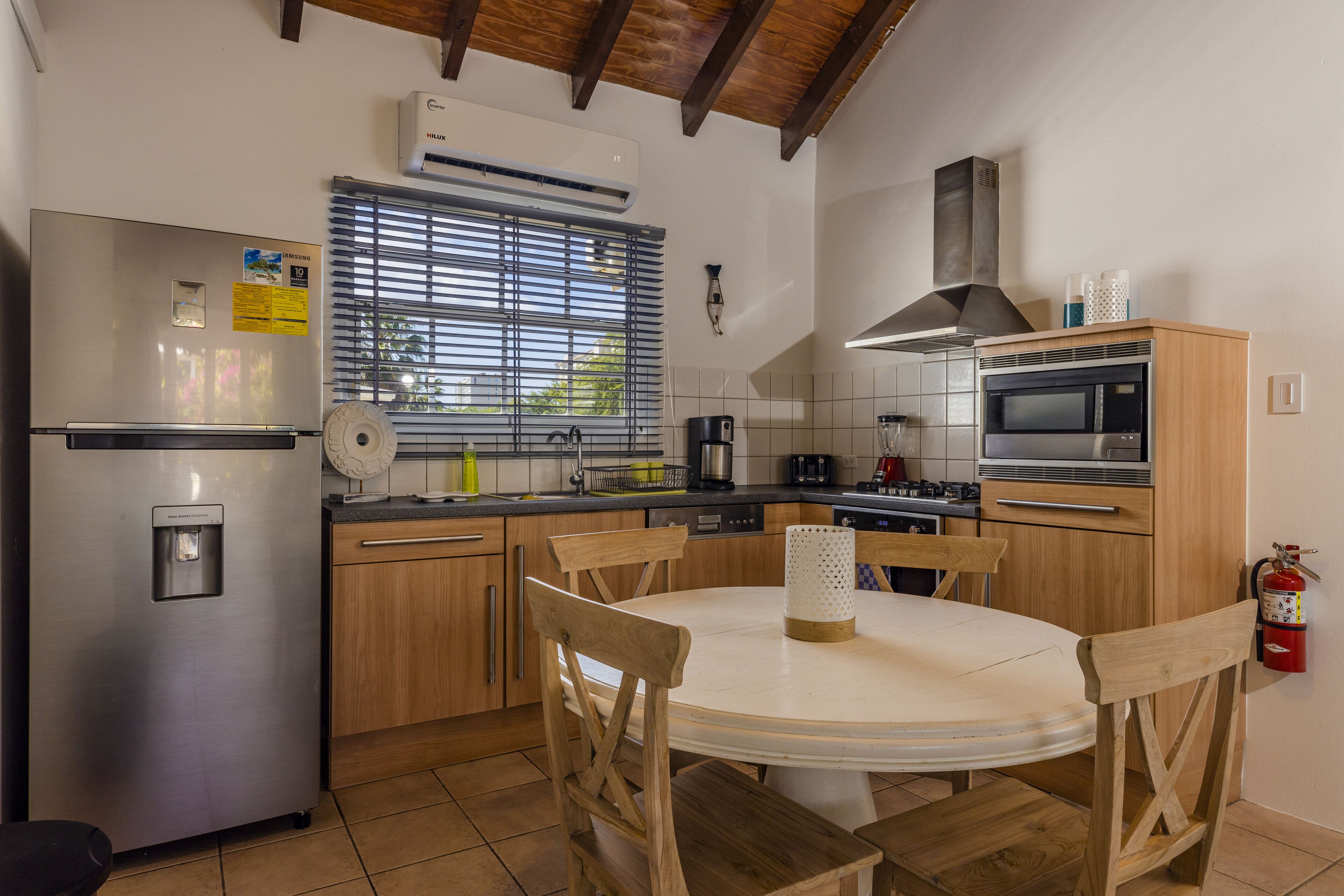 Een keuken met een roestvrijstalen koelkast, houten kastjes, een ronde eettafel en een raam met jaloezieën.
