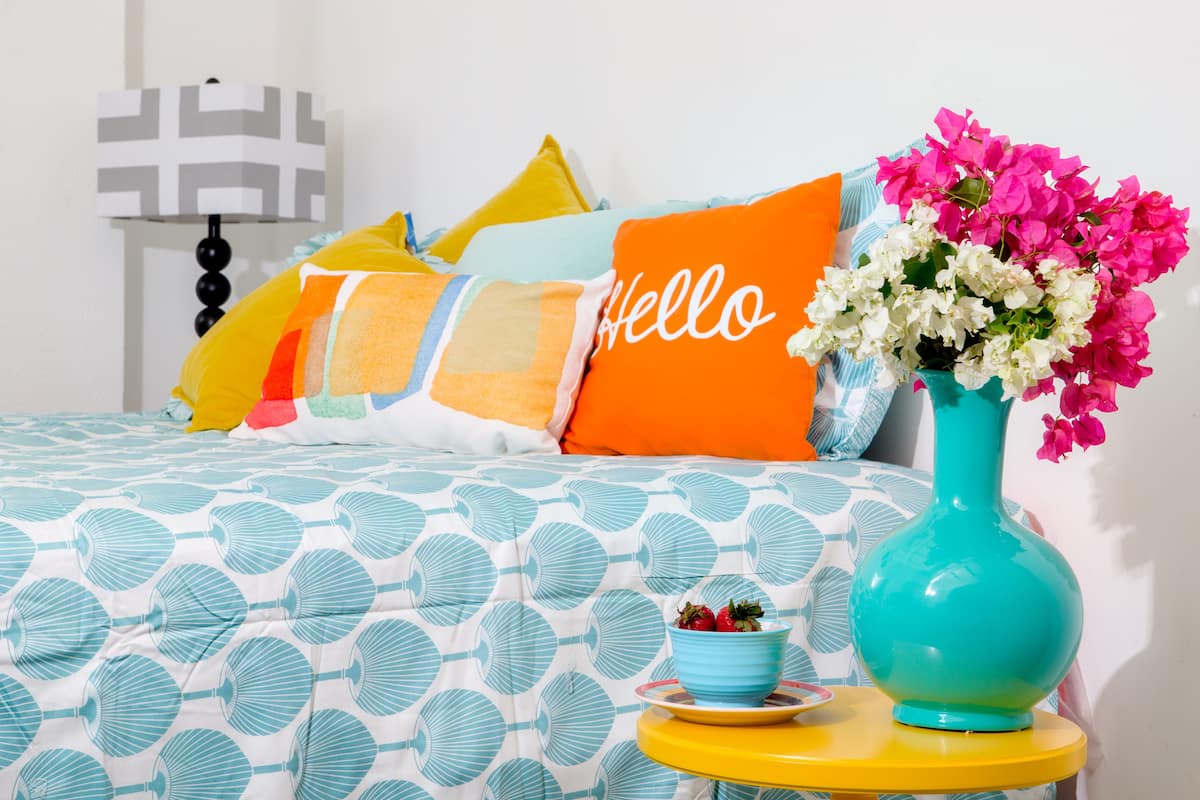 A bed with a patterned bedspread, a vase with flowers, and a bedside table with a bowl of fruit.