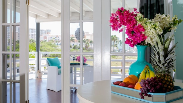 Una habitación con una mesa blanca, un florero turquesa con flores y un frutero.